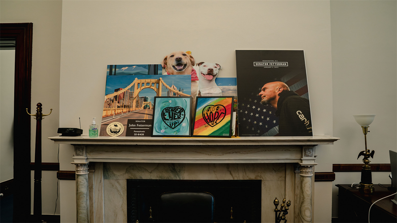 Paintings with uplifting phrases, cut out posters of dogs and posters depicting Senator John Fetterman sit atop a marble fireplace mantel in Fetterman's office