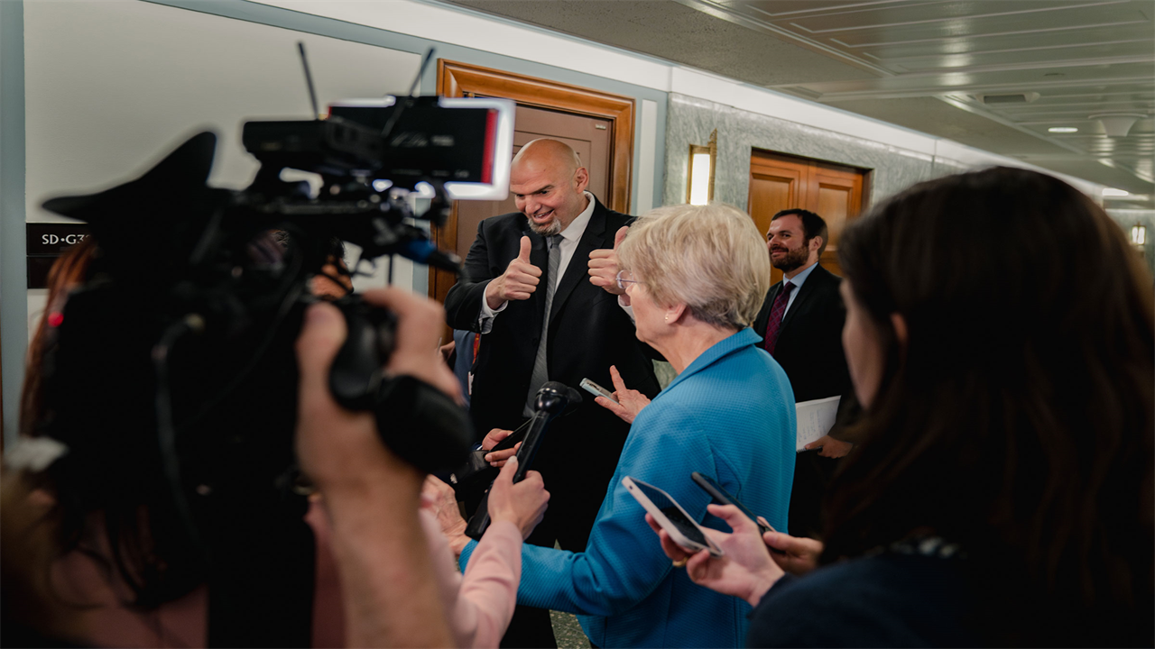 Senator John Fetterman hold both of his thumbs in the air as he walks by Senator Elizabeth Warren in a hallway of Capitol Hill. Warren, in a blue blazer is standing in front of cameras and reporters doing interviews